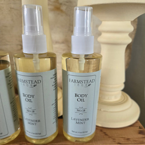 Two Farmstead 1865 Lavender Mint body oil spray bottles on a wooden surface, with a cream pedestal and neutral background.